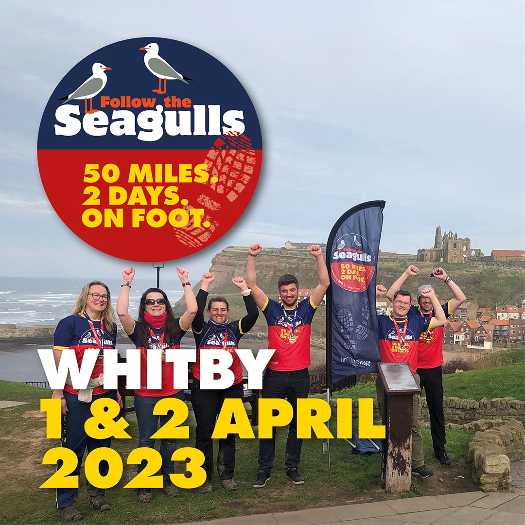 Text reads: Whitby 1 and 2 April 2023. Image description: group of charity walkers wearing follow the seagulls t-shirts are stood cheering on cliff top with Whitby castle behind them.