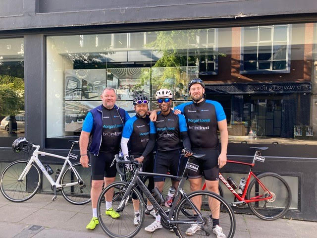 Group of cyclists from Morgan Lovell fundraise for brainstrust. Image shows team of 4 men in cycling gear, they stand arm in arm in front of their bikes smiling. 
